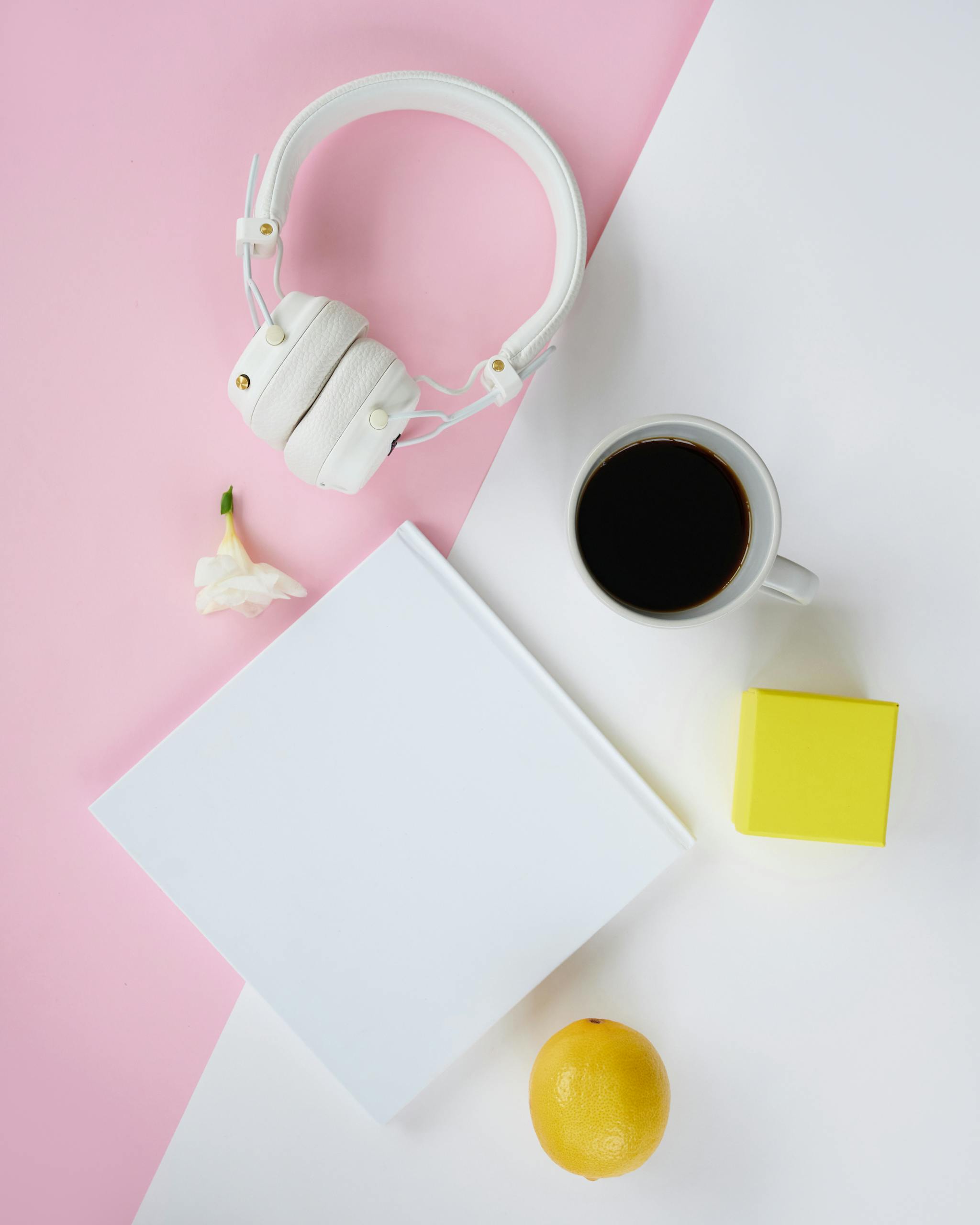 Aesthetic flat lay with headphones, coffee, book, and minimalist decor on a pink and white background.